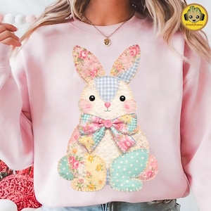Puede incluir: Una sudadera rosa claro con un diseño de conejo de retazos. El conejo tiene patrones florales y de cuadros en rosa, azul y amarillo. Tiene una pajarita y detalles de botones. La sudadera es de estilo casual y cómodo.