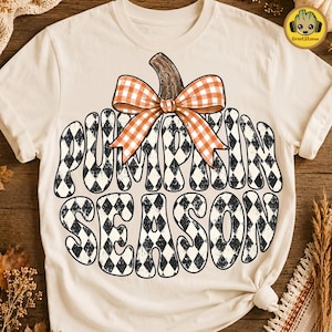 Könnte beinhalten: Ein cremefarbenes T-Shirt mit einer großen Grafik in Kürbisform, die mit einem schwarz-weißen Rautenmuster gefüllt ist. Der Text "PUMPKIN SEASON" ist in einer geschwungenen Schrift im Kürbis abgebildet, gekrönt von einer orange-weißen Vichy-Schleife.