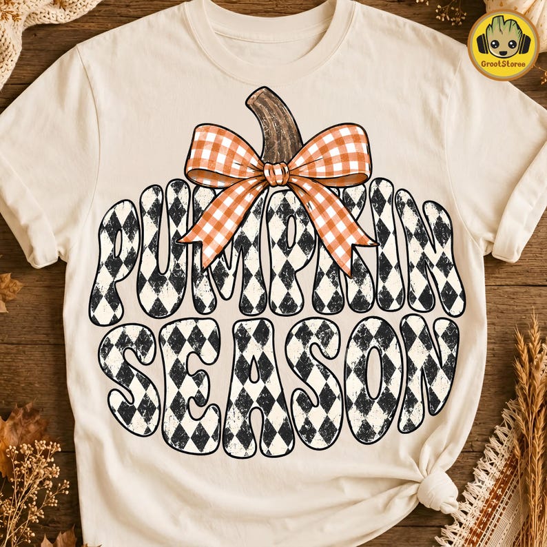 May include: A cream-colored t-shirt featuring a large graphic of a pumpkin shape filled with a black and white diamond pattern. The text "PUMPKIN SEASON" is written in a wavy font within the pumpkin, topped with an orange and white gingham bow.