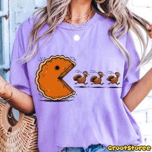 May include: A light purple t-shirt featuring a cartoon design of a pie slice chasing three turkeys. The pie slice is orange with a brown crust, and the turkeys are brown with red wattles. The text "GrootStoree" is in the bottom right corner.