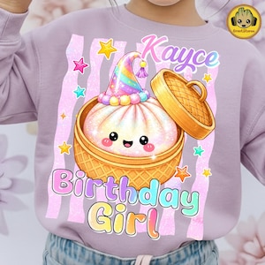 May include: A light purple sweatshirt featuring a graphic of a smiling steamed bun in a bamboo steamer basket, wearing a rainbow party hat. The text reads Kayce Birthday Girl, surrounded by colourful stars and vertical glittery pink stripes.