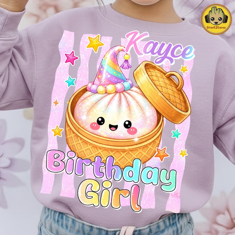 May include: A light purple sweatshirt featuring a graphic of a smiling steamed bun in a bamboo steamer basket, wearing a rainbow party hat. The text reads Kayce Birthday Girl, surrounded by colourful stars and vertical glittery pink stripes.