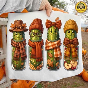 May include: A white t-shirt featuring a graphic print of four green pickles in glass jars. Each pickle is dressed for autumn with accessories including a brown hat, glasses, a bow, and plaid scarves, surrounded by fall leaves, acorns, and apple slices.