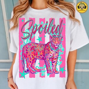 May include: White t-shirt featuring a colorful illustration of a leopard wearing a crown and necklace. The word "Spoiled" is written in cursive above the leopard, set against a pink and white striped background with teal accents. The design is vibrant and eye-catching.
