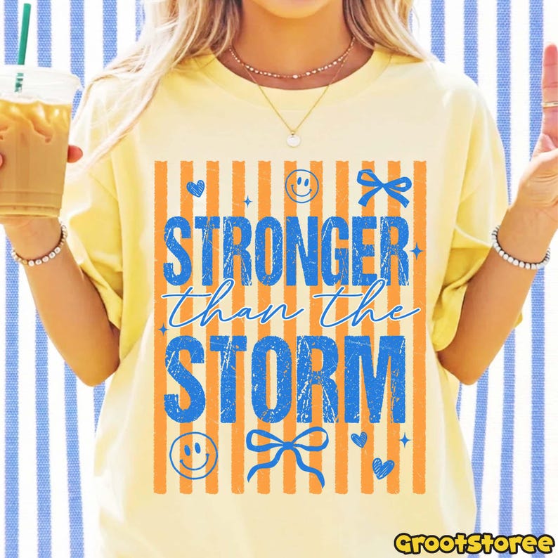 K&ouml;nnte beinhalten: Gelbes T-Shirt mit dem blauen Schriftzug "STRONGER than the STORM", verziert mit orangefarbenen Streifen, Herzen, Schleifen und Smileys. L&auml;ssiger, bequemer Stil.