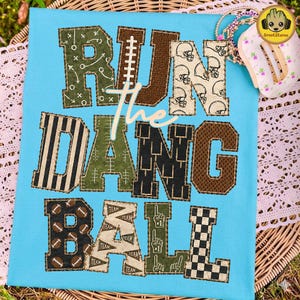 May include: A light blue t-shirt featuring the text RUN THE DANG BALL in large, block-style letters. Each letter displays a unique football-themed pattern, including helmets, laces, goal posts, pennants, foam fingers, and checkered designs.