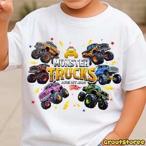 May include: White t-shirt featuring a colorful graphic of monster trucks. The design includes various monster trucks in orange, yellow, red, blue, green, and purple, with the text "MONSTER TRUCKS ARE MY JAM".
