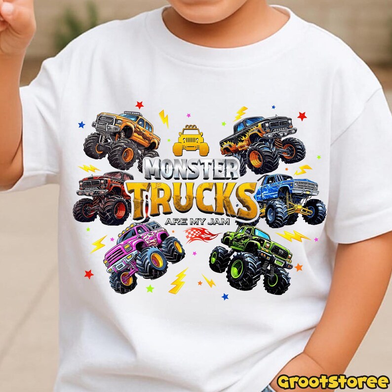 May include: White t-shirt featuring a colorful graphic of monster trucks. The design includes various monster trucks in orange, yellow, red, blue, green, and purple, with the text "MONSTER TRUCKS ARE MY JAM".