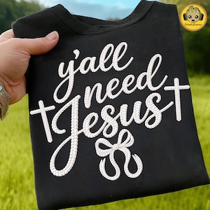 May include: Black crew neck t-shirt with the phrase "y'all need Jesus" in white script. The text includes a cross and a decorative bow. The shirt is held up against a background of green grass and blue sky.