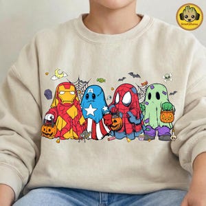 May include: A beige crewneck sweatshirt featuring a graphic print of four characters dressed as ghosts for Halloween. The characters resemble Iron Man, Captain America, Spider-Man, and a green ghost, each holding a small orange jack-o'-lantern bucket.