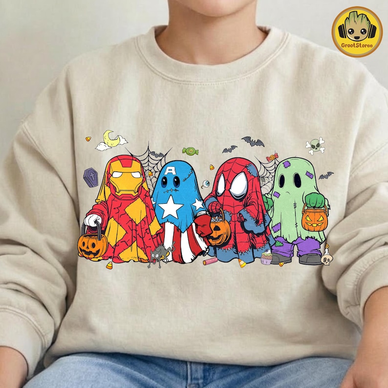 May include: A beige crewneck sweatshirt featuring a graphic print of four characters dressed as ghosts for Halloween. The characters resemble Iron Man, Captain America, Spider-Man, and a green ghost, each holding a small orange jack-o'-lantern bucket.