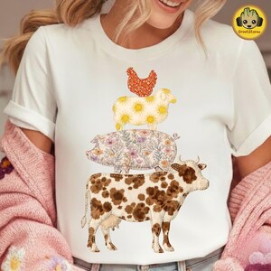以下が含まれることがあります： 白いTシャツ。積み重ねられた農場の動物のデザイン。茶色と白の牛が、花柄の豚、太陽のデザインの羊、赤い模様の鶏を支えています。Tシャツはクルーネックです。