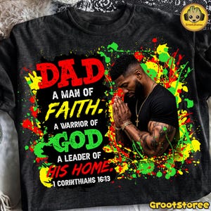 Könnte beinhalten: Ein anthrazitfarbenes T-Shirt mit einer Grafik eines Mannes mit gefalteten Händen im Gebet. Der Text lautet: DAD A MAN OF FAITH, A WARRIOR OF GOD A LEADER OF HIS HOME, 1 CORINTHIANS 16:13. Das Design enthält rote, gelbe und grüne Farbspritzer.