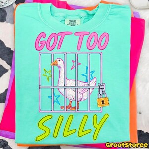 K&ouml;nnte beinhalten: T&uuml;rkisfarbenes Comfort Colors T-Shirt mit einer Grafik einer Gans in einem K&auml;fig, umgeben von Sternen. Der Text "GOT TOO SILLY" ist in Pink und Gelb. Das Shirt liegt gefaltet auf anderen bunten Shirts.