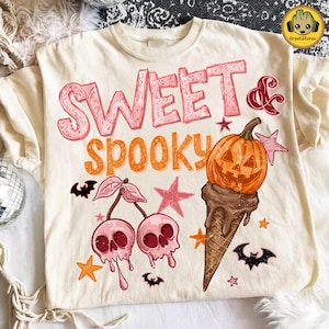 Może przedstawiać: Kremowy t-shirt z napisem SWEET & SPOOKY w kolorze różowym i pomarańczowym. Grafika przedstawia dwie różowe wiśnie w kształcie czaszek z ogonkami, rożek lodowy z dynią i czekoladą oraz kilka małych czarnych nietoperzy i pomarańczowych gwiazdek.