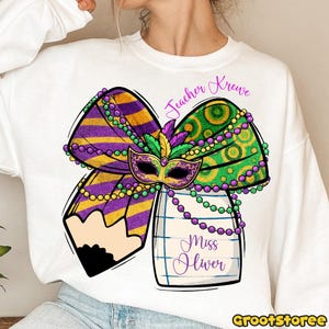 May include: White sweatshirt featuring a colorful Mardi Gras-themed design. The graphic includes a purple and gold bow, a green and gold bow, a mask, and a pencil. The text "Teacher Krewe" and "Miss Oliver" are also included.