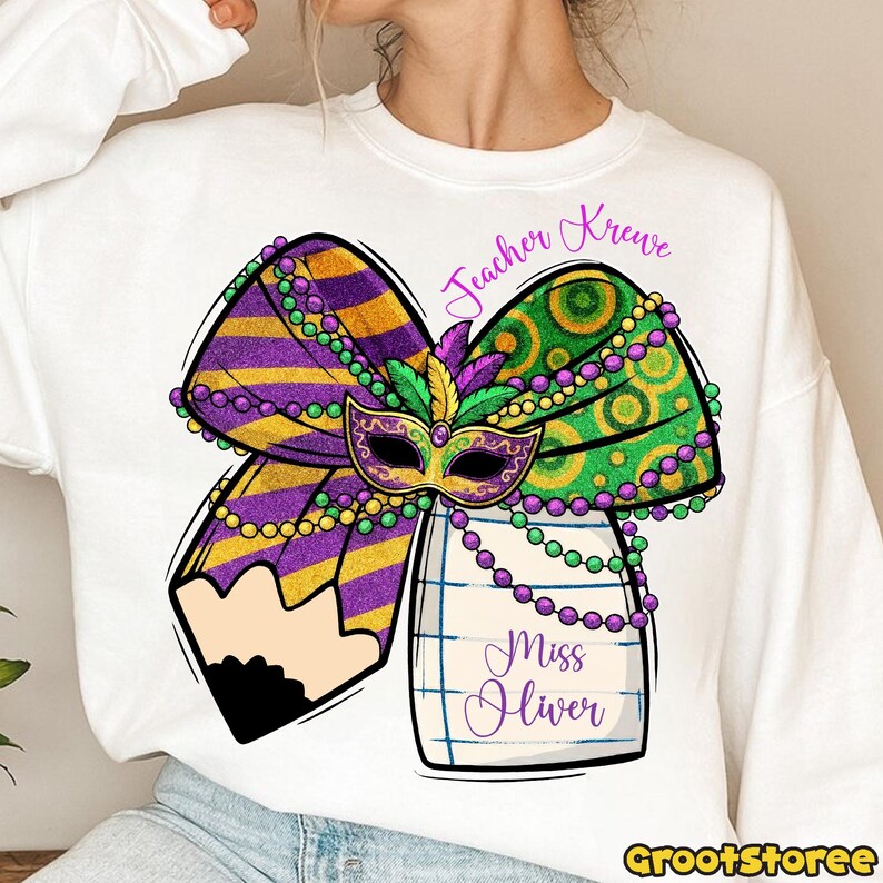 May include: White sweatshirt featuring a colorful Mardi Gras-themed design. The graphic includes a purple and gold bow, a green and gold bow, a mask, and a pencil. The text "Teacher Krewe" and "Miss Oliver" are also included.