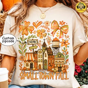 May include: A cream-colored t-shirt featuring a colorful graphic of a small town with buildings, a water tower, and autumn foliage. The design includes a sign with the text 240963, a custom zipcode label, and the text SMALL TOWN FALL at the bottom.