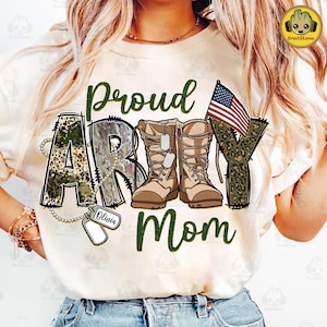 May include: White t-shirt with the text "Proud Army Mom" in green and camouflage print. The word "Army" is decorated with camouflage print and a pair of brown military boots. A small American flag is on the right side of the shirt. A silver chain with a dog tag that says "Olivia" is hanging from the bottom of the "A" in "Army".