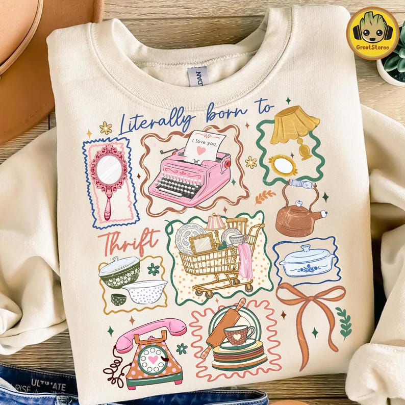 May include: Cream-colored sweatshirt with a whimsical graphic design. The design includes a pink typewriter, a mirror, a shopping cart, and a telephone. The text "Literally born to Thrift" is also featured. The sweatshirt is made of a soft material.
