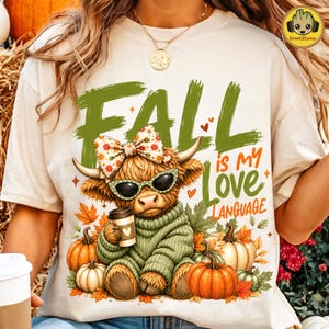 May include: A cream-colored t-shirt featuring a graphic of a Highland cow wearing a floral bow and sunglasses, holding a coffee cup. The text reads FALL is my LOVE LANGUAGE. The design includes orange pumpkins and autumn leaves in the background.