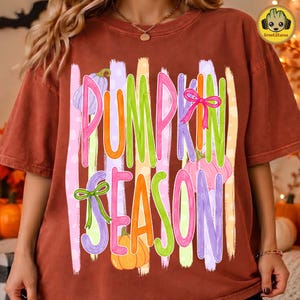 May include: A rust-colored t-shirt featuring a graphic print with vertical pastel stripes and the text PUMPKIN SEASON in colorful, stylized letters. The design includes illustrations of pumpkins in purple, pink, and orange, accented with small green bows.