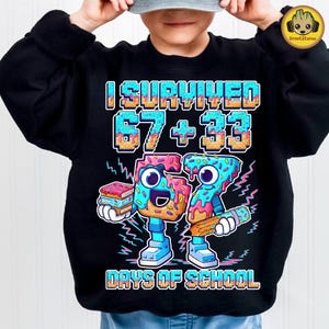 Peut inclure: Sweat-shirt noir avec le texte "I Survived 67 + 33 Days of School". Le graphisme présente deux personnages en forme de beignets tenant des livres et un crayon. Les beignets sont bleus, roses et jaunes.
