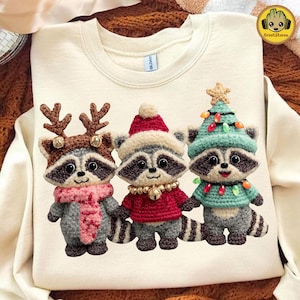 Peut inclure: Sweat-shirt crème avec trois ratons laveurs en crochet vêtus de tenues de Noël. L'un porte des bois de renne et une écharpe rose, un autre un bonnet de Père Noël et un pull rouge, et le troisième un chapeau sapin et un pull vert.