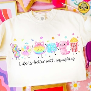 May include: A cream-colored t-shirt featuring a graphic print of six colorful, smiling cartoon characters including a butter stick, paw print, rainbow ball, ice cube, axolotl, and popcorn bucket, with the text Life is better with squishies.