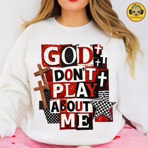 May include: White sweatshirt with the text "GOD DON'T PLAY ABOUT ME" in a bold, layered design. The design includes red and black blocks, wooden crosses, and a checkered star, creating a striking visual statement.