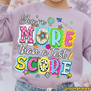 May include: A light purple sweatshirt with the text "You're MORE than a test SCORE" in colorful block letters. The design includes school-themed graphics like a globe, stars, and a pencil. The sweatshirt is suitable for children.