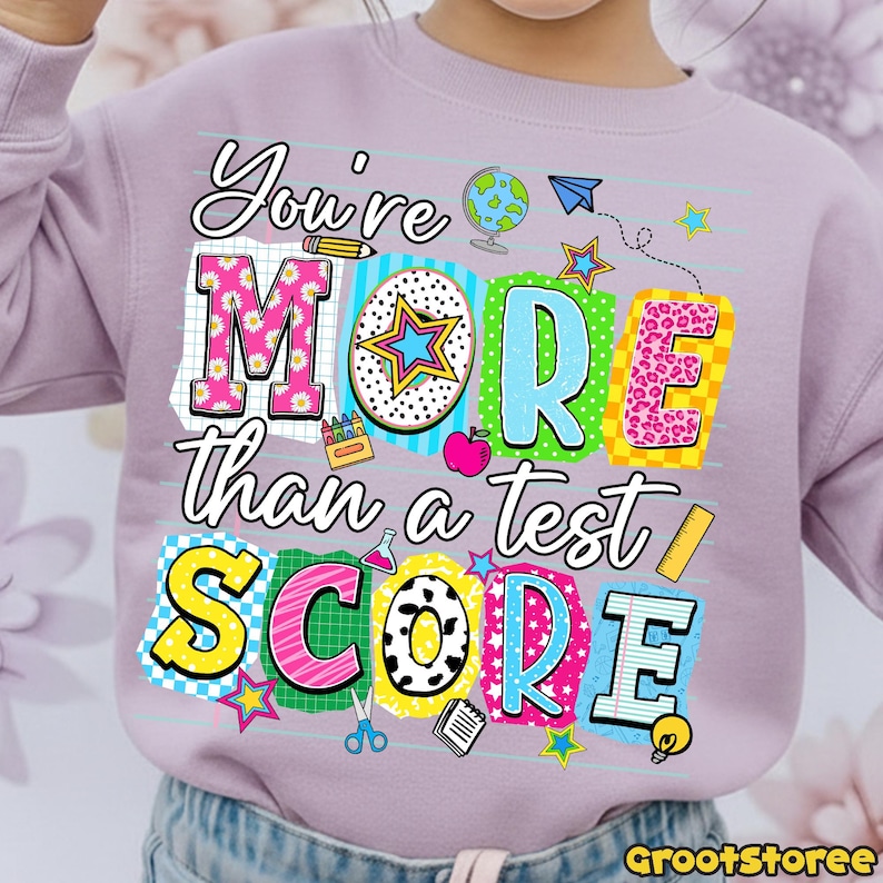 May include: A light purple sweatshirt with the text "You're MORE than a test SCORE" in colorful block letters. The design includes school-themed graphics like a globe, stars, and a pencil. The sweatshirt is suitable for children.