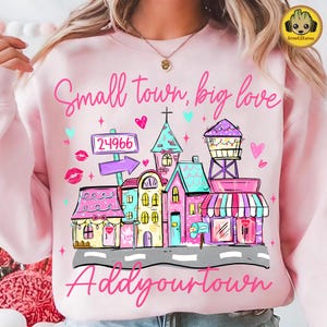 Puede incluir: Sudadera rosa claro con un gráfico de ciudad colorida. El diseño incluye edificios, una torre de agua y los textos "Small town, big love" y "Add your town". El gráfico también presenta corazones y el número 24966 en una señal.