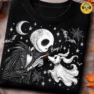 Peut inclure: Un t-shirt noir avec une illustration graphique blanche représentant un personnage squelettique en costume rayé touchant le nez d'un chien fantôme. Le motif inclut un croissant de lune, des étoiles, des toiles d'araignée, des chauves-souris et le mot Boop.