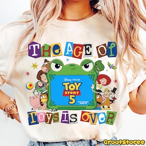 May include: A cream-colored t-shirt featuring a graphic of various Toy Story characters surrounding a green frog-shaped frame. The frame contains the text Disney Pixar Toy Story 5 In Theaters June 19. The text The Age Of Toys Is Over is printed.