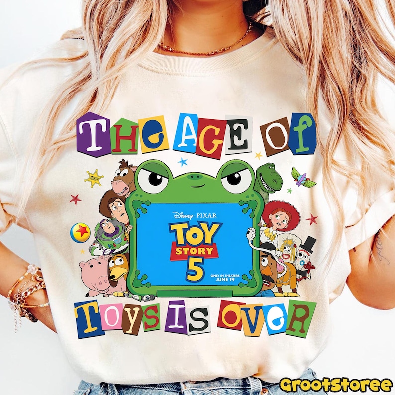 May include: A cream-colored t-shirt featuring a graphic of various Toy Story characters surrounding a green frog-shaped frame. The frame contains the text Disney Pixar Toy Story 5 In Theaters June 19. The text The Age Of Toys Is Over is printed.