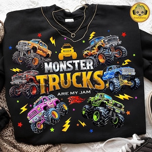 May include: Black sweatshirt featuring a colorful graphic of several monster trucks with large tires. The text "MONSTER TRUCKS ARE MY JAM" is prominently displayed. Lightning bolts and stars surround the trucks, creating a dynamic design.