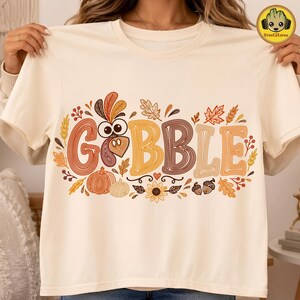 May include: A cream-colored t-shirt featuring the word GOBBLE in large, multi-colored, line-art style letters. The design includes a turkey head replacing the letter O, surrounded by autumn-themed illustrations like pumpkins, leaves, and acorns.