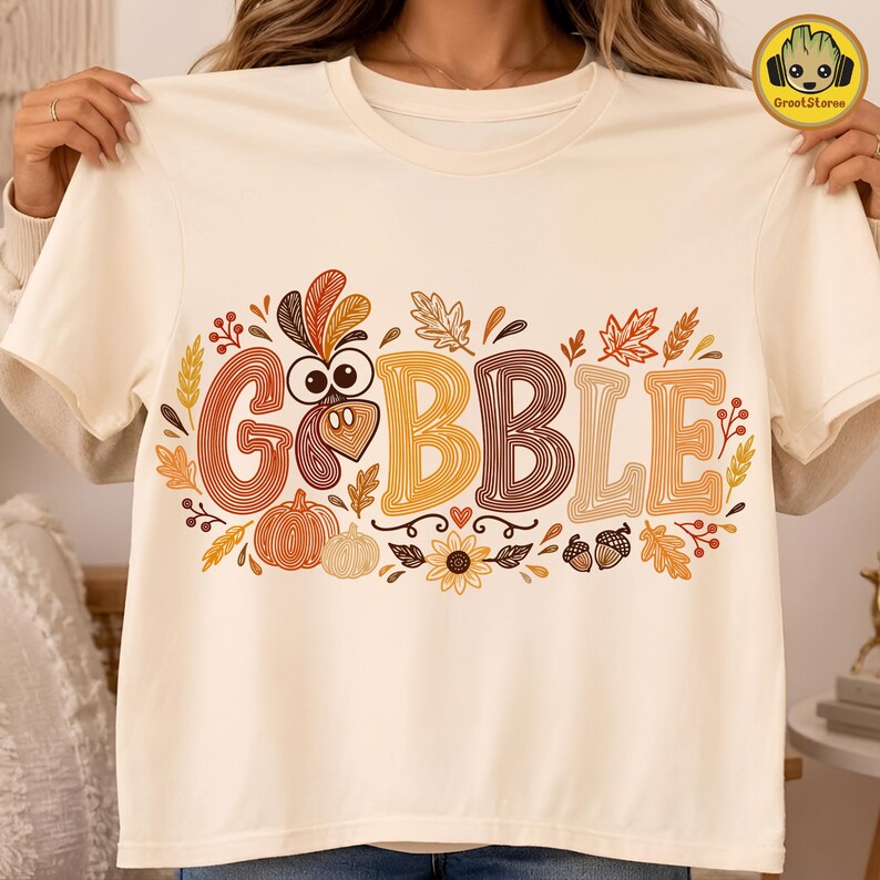May include: A cream-colored t-shirt featuring the word GOBBLE in large, multi-colored, line-art style letters. The design includes a turkey head replacing the letter O, surrounded by autumn-themed illustrations like pumpkins, leaves, and acorns.