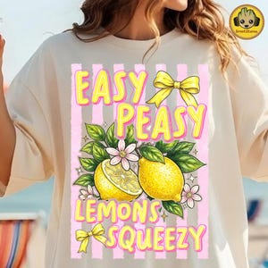 May include: A cream-coloured t-shirt with a graphic design. The design features lemons, flowers, and the text "EASY PEASY LEMONS SQUEEZY" in yellow and pink. The design includes yellow bows and a pink striped background.