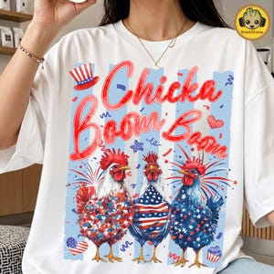 Può includere: T-shirt bianca con un design patriottico con tre galli con creste rosse e motivi a bandiera americana. Il testo "Chicka Boom Boom" è in corsivo rosso con accenti blu, insieme a fuochi d'artificio e stelle.