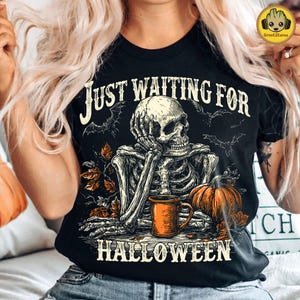 May include: Black t-shirt with a Halloween graphic. The design features a skeleton resting its head on its hand, with a pumpkin and a mug. The text reads "Just Waiting For Halloween" with bats and leaves.