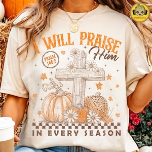 Op de afbeelding: Een crèmekleurig t-shirt met een grafische print met de tekst I WILL PRAISE HIM en IN EVERY SEASON. Het ontwerp bevat een houten kruis, pompoenen, madeliefjes, herfstbladeren en de bijbelverwijzing PSALM 146:2 in een cirkelvormig kader.
