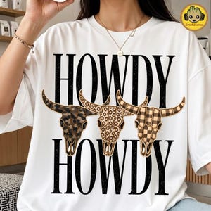 May include: A white t-shirt featuring the black text HOWDY printed twice in a distressed font. Between the text lines are three bull skulls with different patterns: one with dark camouflage, one with leopard print, and one with a brown plaid pattern.