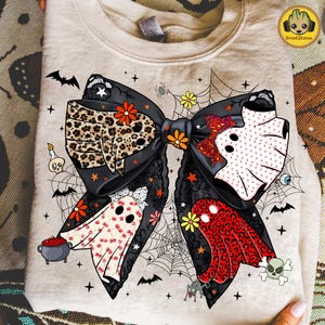 May include: Beige sweatshirt featuring a large bow design with Halloween-themed elements. The bow has a leopard print section, ghost figures, and spiderwebs. Additional graphics include bats, stars, and a skull. The design incorporates orange, red, and black colors.