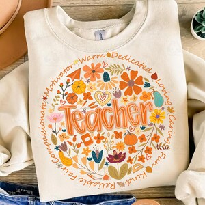 May include: Cream-colored sweatshirt with a floral design and the word "Teacher" in orange. The design includes flowers, pumpkins, and the words "Motivator," "Warm," "Dedicated," "Loving," "Caring," "Fun," "Kind," "Reliable," and "Fair."