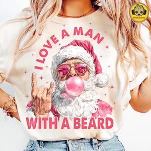 May include: Cream-colored t-shirt with a graphic of Santa Claus wearing pink sunglasses and blowing a bubblegum bubble. The text "I LOVE A MAN WITH A BEARD" is printed in pink. Pink stars are scattered around the graphic.