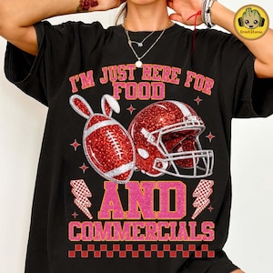 May include: Black t-shirt with a graphic design featuring a red American football with bunny ears, a red helmet, and the text "I'M JUST HERE FOR FOOD AND COMMERCIALS" in pink and white.