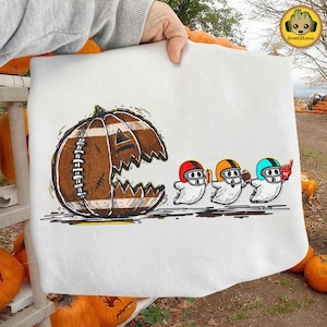 Pode incluir: Camisola branca com um design de Halloween. Uma abóbora em forma de bola de futebol com uma boca irregular está à esquerda. Três fantasmas de desenho animado usando capacetes de futebol estão à direita.