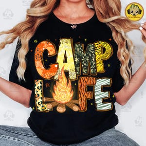 Peut inclure: T-shirt noir avec le texte "Camp Life" en lettres colorées et à motifs. Les lettres sont composées de différents imprimés animaliers, dont léopard, zèbre et tigre. Un feu de camp est représenté au centre du texte.